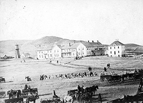 A black and white photo featuring wooden buildings along a barren plain with the rise of a hill in the background and a line of people, mostly women dressed in long skirts, along a pathway in the bottom center.  Two unhitched carriages and 2 men driving a carriage with two horses are in the foreground. 